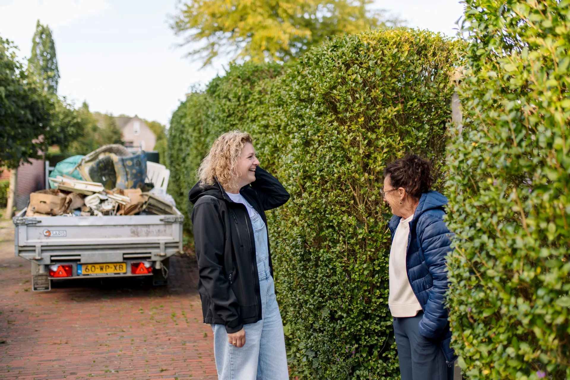 Twee mensen voeren een gesprek naast een heg met een vrachtwagen op de achtergrond.